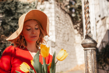 A  beautiful woman dressed in a fashion  red dress, with a bouquet of  yellow flowers in her hands walking along the old town with ancient buildings of the last century