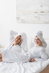 young and pretty interracial women in white soft robes and towels smiling at camera while resting on bed.
