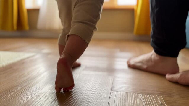 baby newborn first steps. mother teaches daughter baby to take the first steps with bare feet on the floor. baby walks barefoot on the floor at home learning. baby newborn first steps indoors light