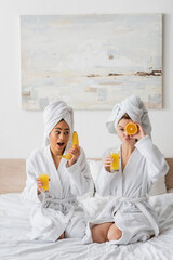 young woman pouting lips and covering eye with ripe orange near african american woman with banana and orange juice.