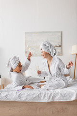 Obraz na płótnie Canvas excited african american woman toasting with champagne near happy friend in white robe and towel in bedroom.
