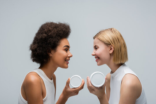 side view of multiethnic blonde and brunette women holding cosmetic cream and smiling at each other isolated on grey.