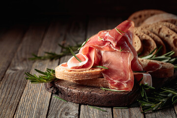 Prosciutto with bread and rosemary on an old wooden table. - obrazy, fototapety, plakaty