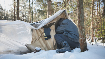 A homeless man climbs in and out of a tent in the woods in winter. - obrazy, fototapety, plakaty