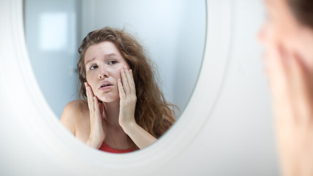 Anxious young woman look in the mirror worried about wrinkle or acne on unhealthy skin, upset unhappy millennial female examine squeeze pimple on face in the bedroom, cosmetology, skincare concept