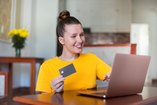 Portrait of happy girl, young positive pretty woman paying, holding credit bank card with phone, laptop. Online shopping at home in living room. Bank application, app.