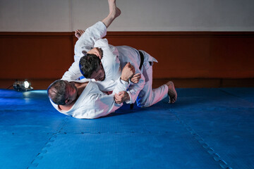 Two latin men training martial arts with kimonos, one black belt and one white belt, one trying to perform a triangle on the teacher.