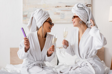happy interracial women in terry robes and stylish sunglasses holding champagne and looking at each other in bedroom.