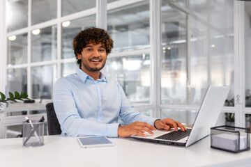 Portrait of young successful contemporary businessman inside office, man at workplace smiling and looking at camera, young hispanic businessman sitting at desk using laptop.