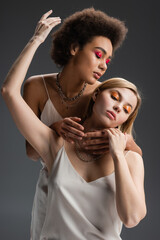 sensual interracial models in white strap dresses and colorful makeup posing isolated on grey.