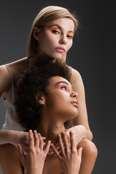 blonde model with orange eye shadows embracing brunette african american woman while looking at camera isolated on grey.