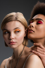blonde woman with orange eye shadows looking away near sensual african american model posing with...