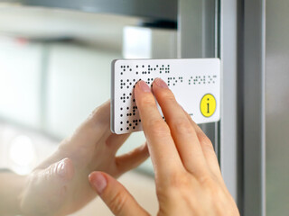 Person with blindness touches and reads with his hands the Braille text plate on the door of a...