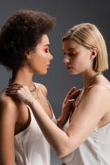 side view of young multiethnic women in strap dresses and silver necklaces touching each other isolated on grey.