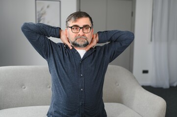 Obraz premium Neck Pain. Portrait Of Exhausted Mature Man Massaging Aching Neck Suffering From Ache Sitting On Couch At Home In Living Room. Osteoarthritis, Older People Health Problem, Sedentary Lifestyle Concept