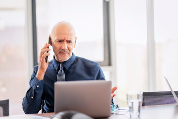 Overwhelmed businesswoman standing in a modern office and using a mobile phone an laptop for work