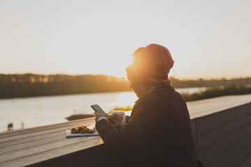 Muslim woman in hijab using smartphone outdoor tasty nuggets and coffee background, close up. Street food and picnic in spring morning