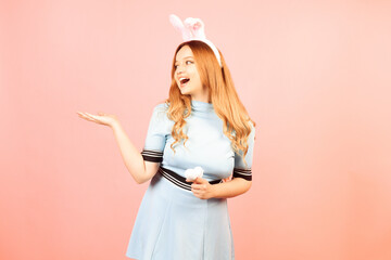A girl with red hair in a blue dress and easter bunny ears on a pink background. A beautiful woman...