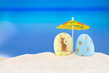 Yellow and blue eggs under umbrella on sandy sea or ocean beach..Creative couple tourist. Happy Easter holiday card on hot country. Travel, spring, summer background. Copy space