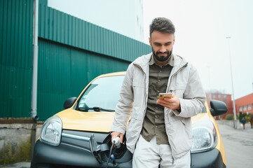 bearded caucasian man standing near an electric car that is charging and making time adjustments on a smartphone.