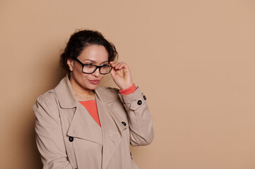 Confident Latin American middle-aged business woman in beige casual coat, putting on stylish specs - eyeglasses, squinting, looking away, standing isolated on beige background. Vision problems concept