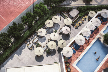 Top view from a drone of an outdoor bar with sunshades creating a beautiful landscape, adjacently a lounge and pool area with people resting, bordering the terrace trees hide the clay tennis court
