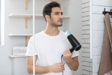 Young handsome man standing in front of the mirror in the bathroom and holding hair dryer. Morning routine and making hair style concept - obrazy, fototapety, plakaty