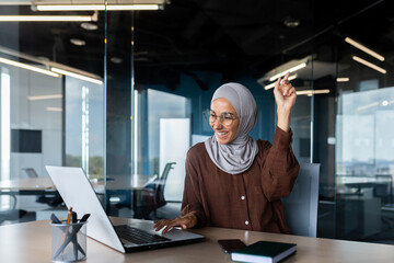 Portrait of happy smiling and successful muslim at work in office, businesswoman with laptop holding hands up gesture of success and triumph, woman in hijab received online victory notification.