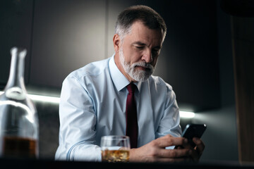 Mature businessman with glass of whiskey using smartphone while sitting in the kitchen.