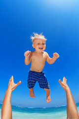Fototapeta na wymiar Family swimming fun on white sand sea beach and blue sky - father hands tossing up baby boy into mid air and catching. Child outdoor activity, active lifestyle on summer vacation in tropical island.