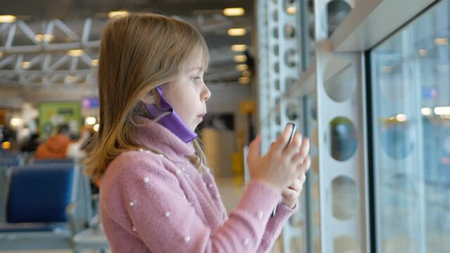 Child traveler take picture for blog in airport look in window. Little girl with face mask use smartphone