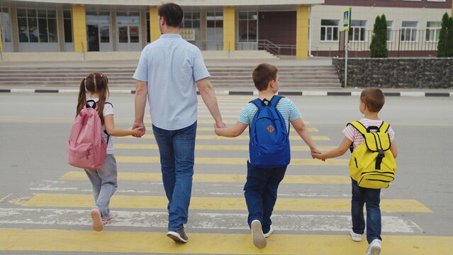 father leads child school holding his hand. dad takes children across road by hand. pedestrian crossing school building. happy family concept. children zebra crossing. parent leads boy girl sister