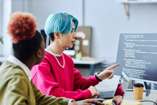 Gen Z creative team working on software programming in office, focus on young Asian man with colored hair pointing at computer screen