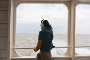 Young latin man wearing headphones on the aft deck of a catamaran while looking at the turbulent water.