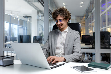 Cheerful and successful businessman inside office working with laptop, bearded man smiling and typing on laptop keyboard, programmer coding software in modern room.
