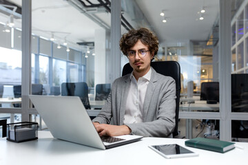 Fototapeta premium Portrait of thinking serious businessman inside office, bearded man looking into camera focused in glasses, worker in business clothes sitting at workplace using laptop at work.