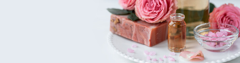Aromatherapy. Pure organic essential rose oil concept. Elixir with plant based floral herbal ingredients. Pink flowers extract. Spa atmosphere with candle, towel. White background. Banner copy space