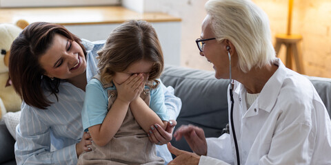 Kind female senior paediatrician doctor visiting his patient at home, examining little girl sitting on mother's lap, writing prescription. Concept of kid's health check. Successful recovery. Banner