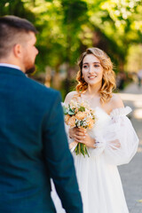 a bride in a delicate dress with a bouquet stands in front of the groom.