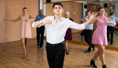 Fototapeta na wymiar Dynamic young man practicing ballroom dance pose in training hall with other persons during dancing-classes