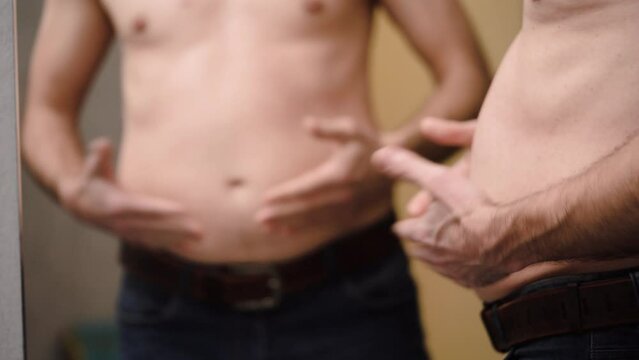 Overweight man holds folds of fat with his hands, looking at his reflection in mirror. 