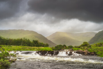 Bundoragha River with small waterfalls and Glenummera and Glencullin mountain ranges illuminated by sunlight in background, County Mayo, Ireland