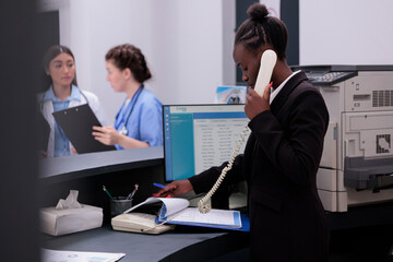 Worker talking at landline phone with remote patient while checking medical appointment papers, standing at hospital reception counter. Health care support service and concept