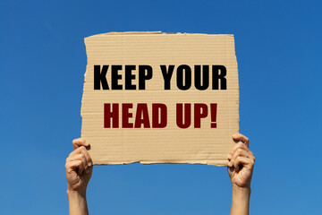 Keep your head up text on box paper held by 2 hands with isolated blue sky background. It is for...