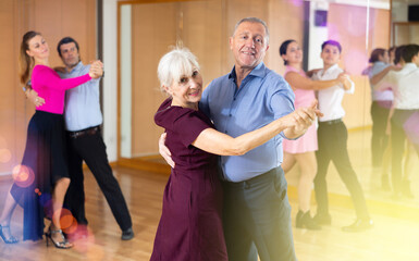 Smiling senior woman and man dancing slow ballroom dance during group class in choreography studio - obrazy, fototapety, plakaty