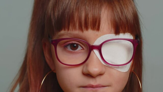 Close-up portrait sick ill preteen girl having protective bandage plaster patch scratch on one eye. Injury, trauma, blind. Child kid wearing glasses at gray background. Medical eyesight vision therapy