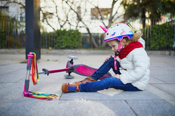 Preschooler girl in unicorn helmet sitting on the ground after she fell while riding her scooter in park on a spring day