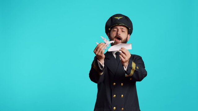 Airline pilot playing with paper and mini airplane, having fun with origami and small artificial aircraft in studio. Young male aviator wearing flying uniform, working on commercial flights.