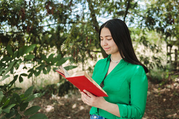 Fototapeta premium Young woman with a green blouse, enjoying reading a book in the shade of trees.