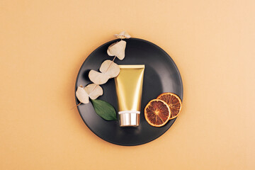 Golden cream tube and dry orange slices, eucalyptus leaves on black plate on beige background. Top view, flat lay, mockup